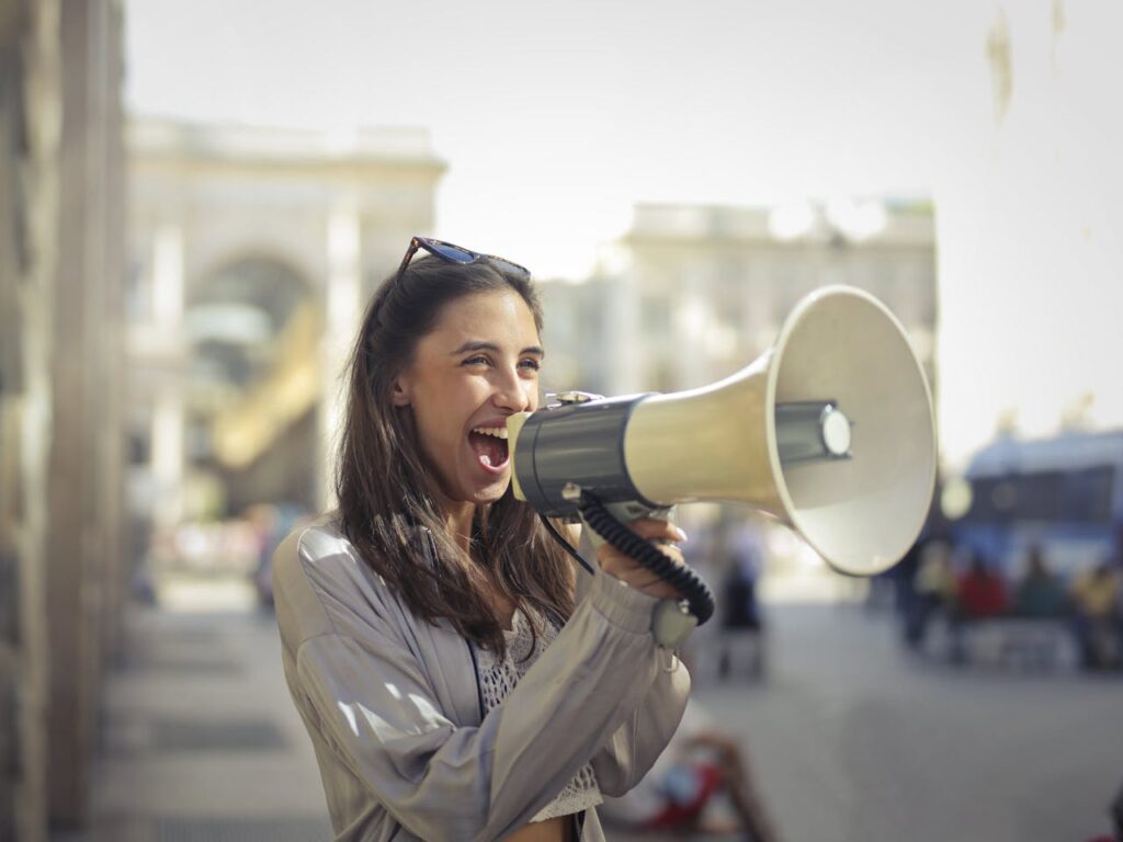 pexels-photo-3761509 Cheerful young woman in a casual outfit shouting into a megaphone on a sunny day.